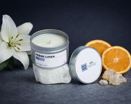 Candle in a tin with 'Fresh Linen' label, surrounded by flowers and oranges on a dark background