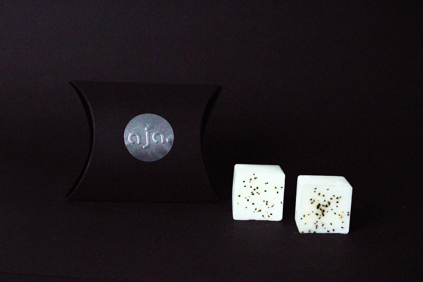 A black pillow box with a circular logo beside two lavender botanical wax melt cubes on a dark background, conveying a minimalist and elegant vibe.
