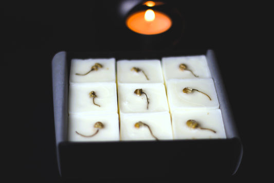 Close-up of square wax melts with lavender pieces in a black box, set against a black background. A small, lit candle flickers softly nearby, adding warmth.