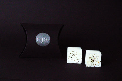 A black pillow box with a circular logo beside two  lavender botanical wax melt cubes on a dark background, conveying a minimalist and elegant vibe.