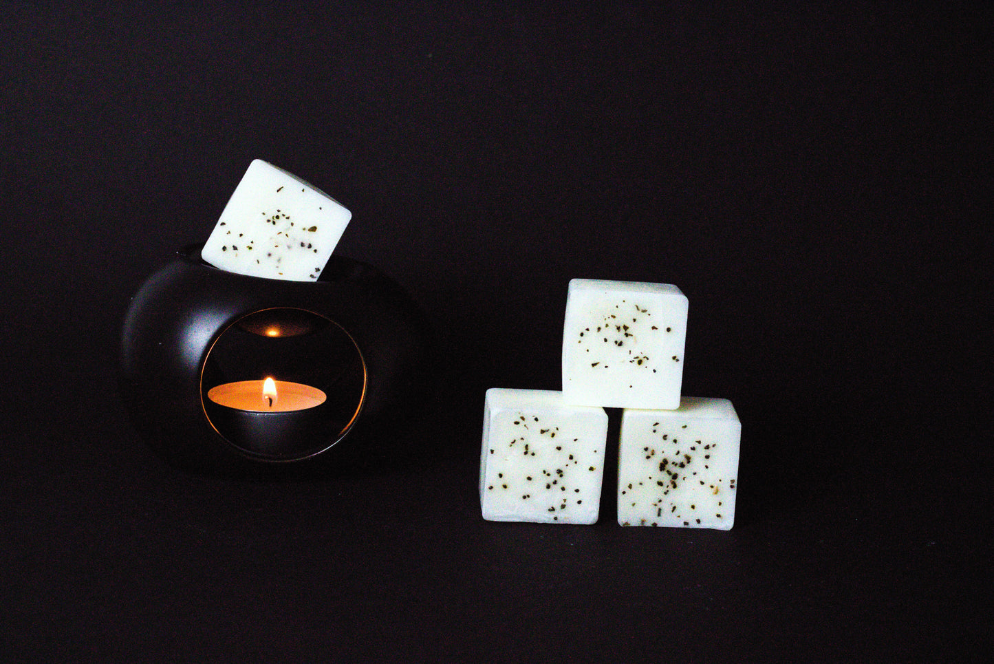 Black candle holder with a lit tealight creates a warm glow. Beside it, three white wax cubes with dried chamomile flower embedded on it