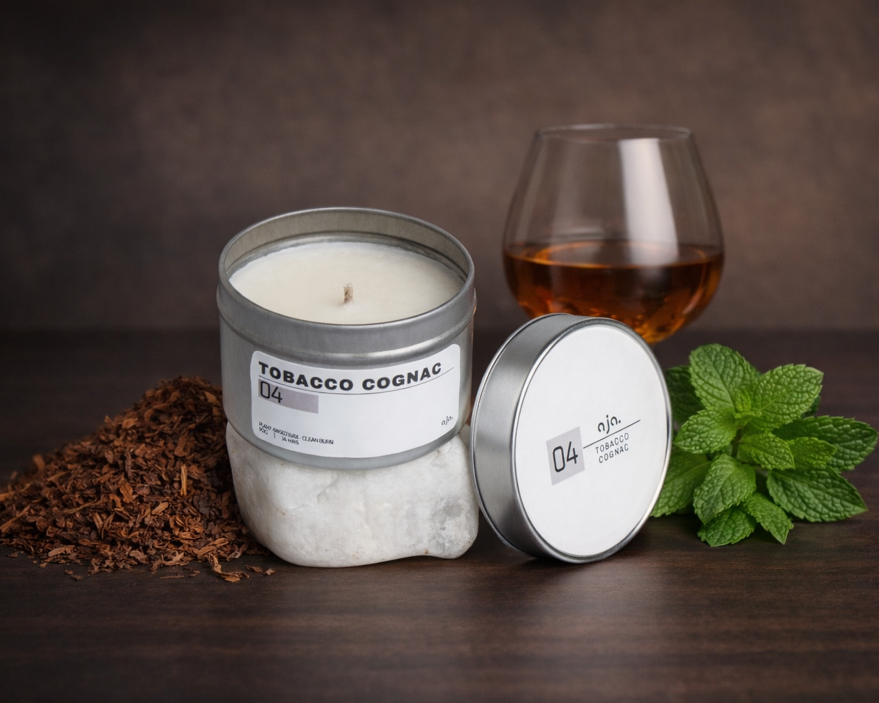Candle labeled 'Tobacco Cognac' with a glass of liquid and mint leaves on a dark surface.