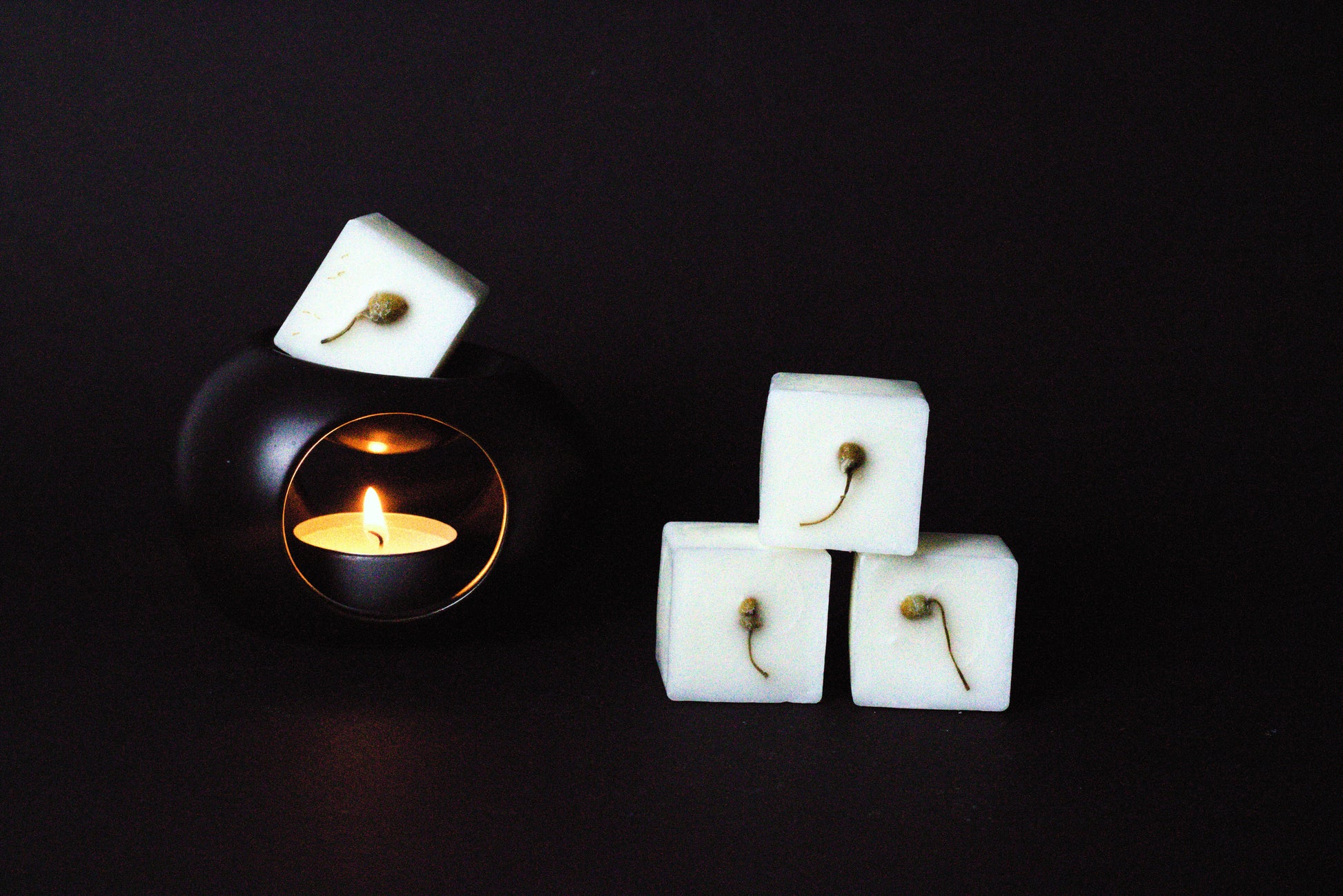 Black candle holder with a lit tealight creates a warm glow. Beside it, three white wax cubes with dried chamomile flower embedded on it