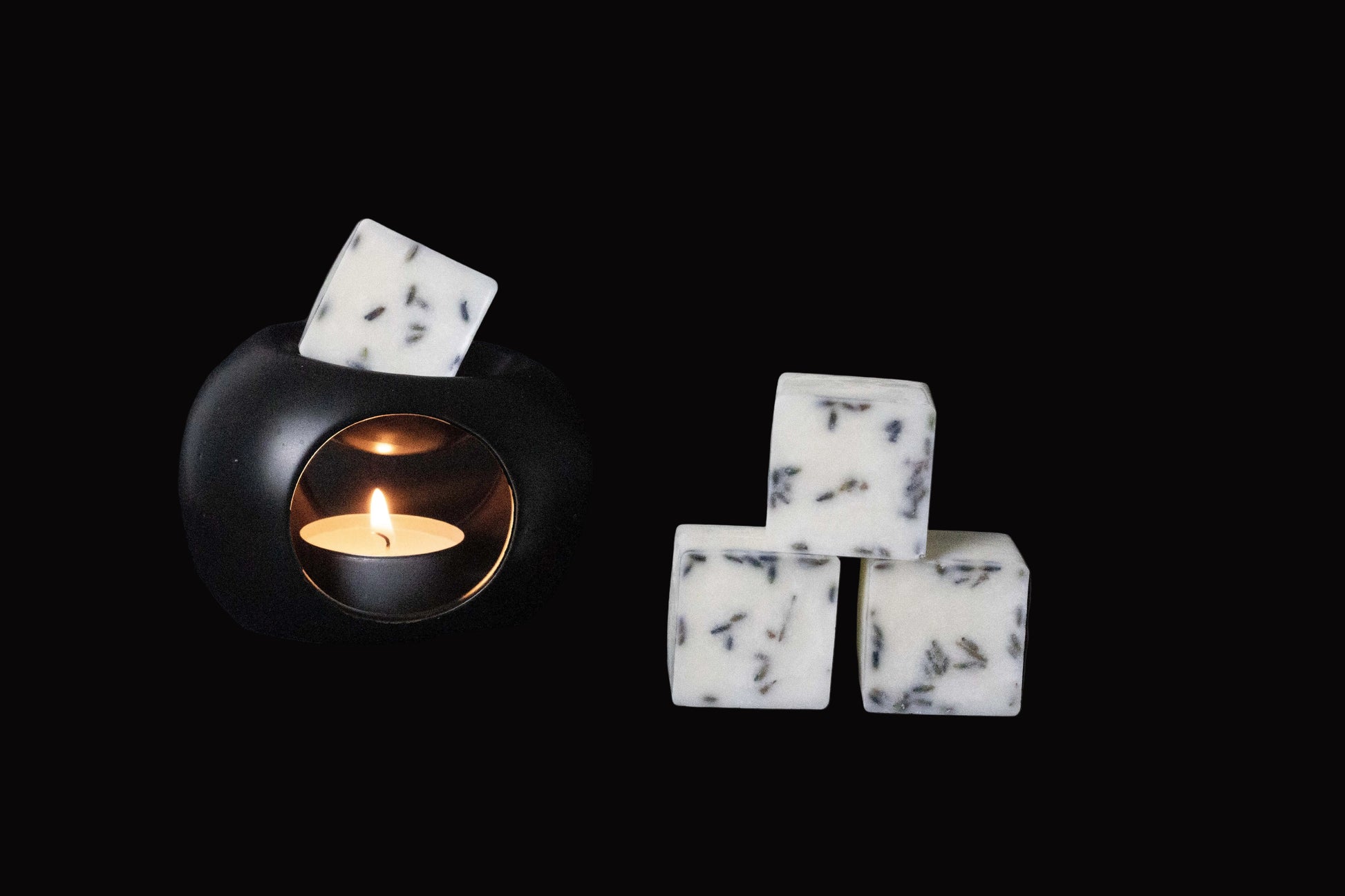Black candle holder with a lit tealight creates a warm glow. Beside it, three white wax cubes with dark flecks are stacked on a dark surface.