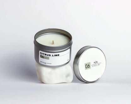 A white candle in a metal tin labeled "Citrus Lime" sits upright. Its matching lid rests beside it. 