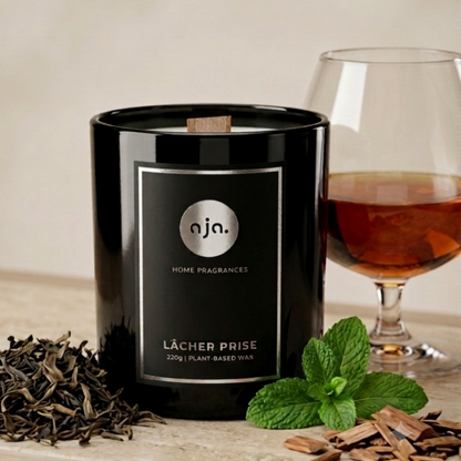 Black candle labeled 'aja Home Fragrances' with a glass of cognac, tobacco leaves and mint leaves on a wooden surface.