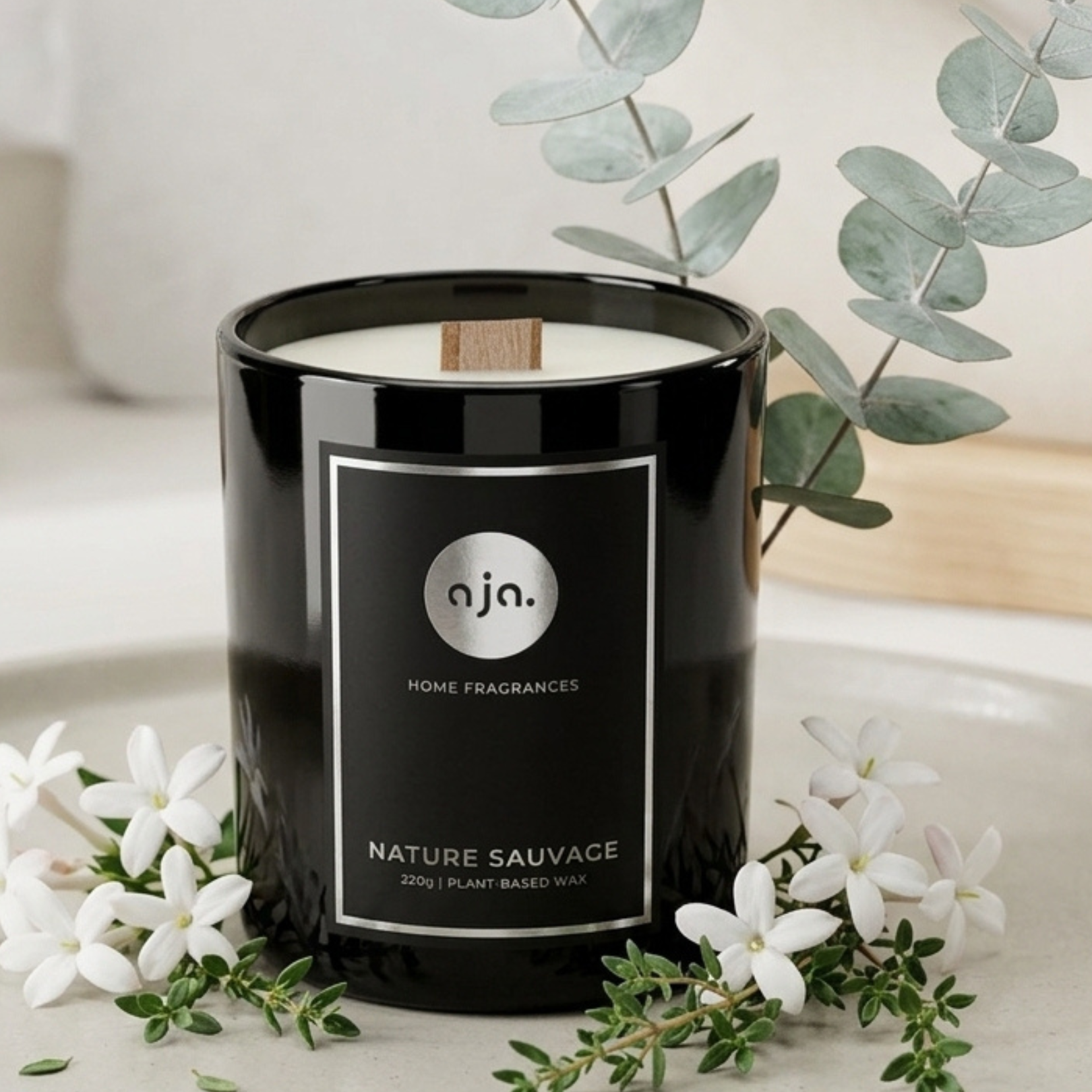 Black candle labeled 'aja Home Fragrances' with eucalyptus and white flowers on a light surface.