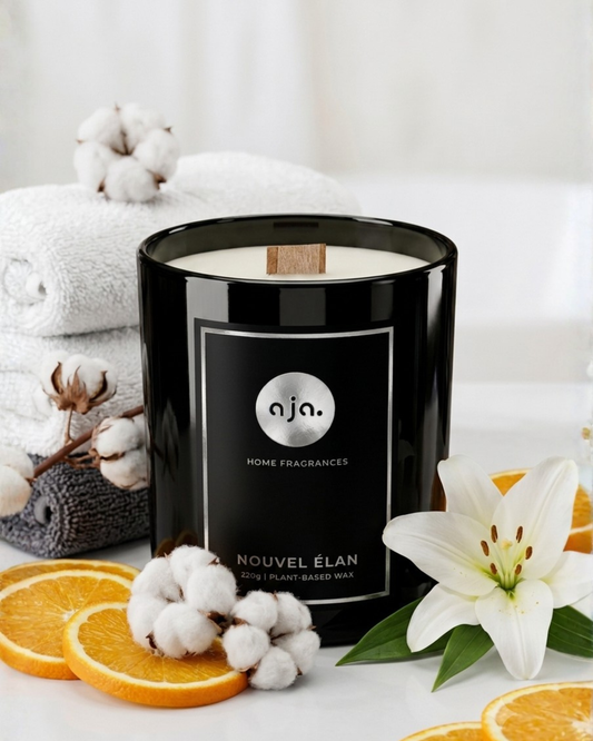 Black candle with 'aja' branding on a white surface with cotton, oranges, and flowers.