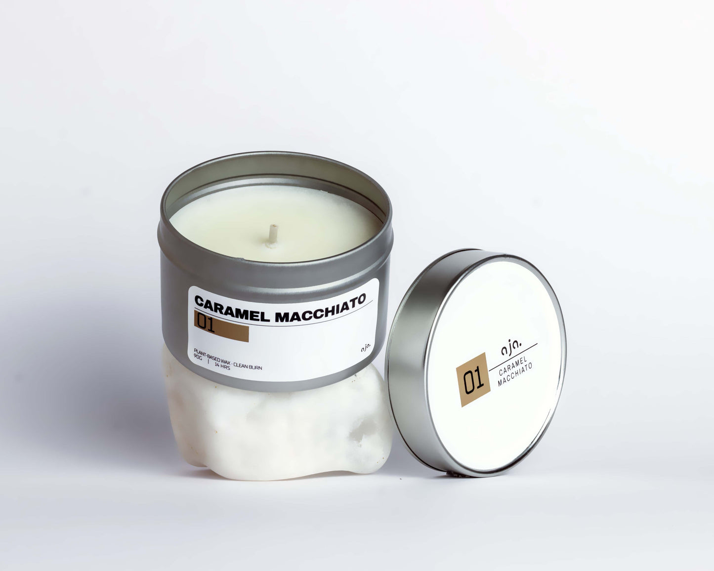 A caramel macchiato-scented candle in a silver tin is open on a white surface. The lid, resting beside it, is labeled "01" with a minimalist design.