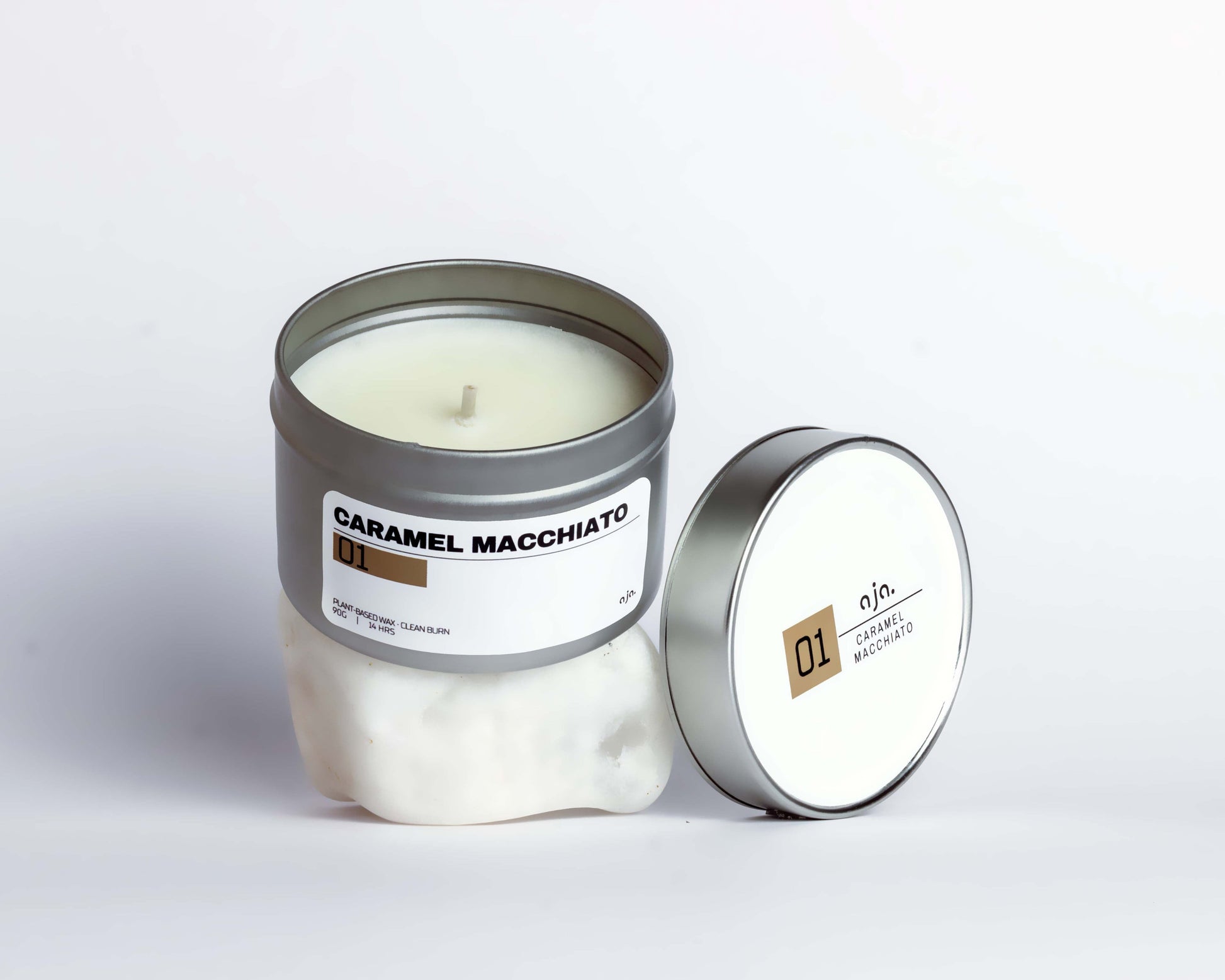 A caramel macchiato-scented candle in a silver tin is open on a white surface. The lid, resting beside it, is labeled "01" with a minimalist design.