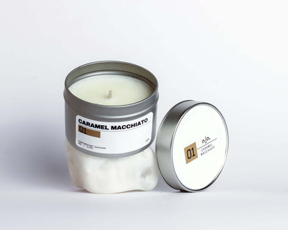 A caramel macchiato-scented candle in a silver tin is open on a white surface. The lid, resting beside it, is labeled "01" with a minimalist design.