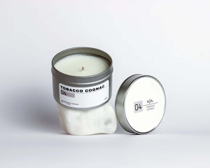 A white candle in a metal tin labeled "Tobacco Cognac" sits upright. Its matching lid rests beside it. 