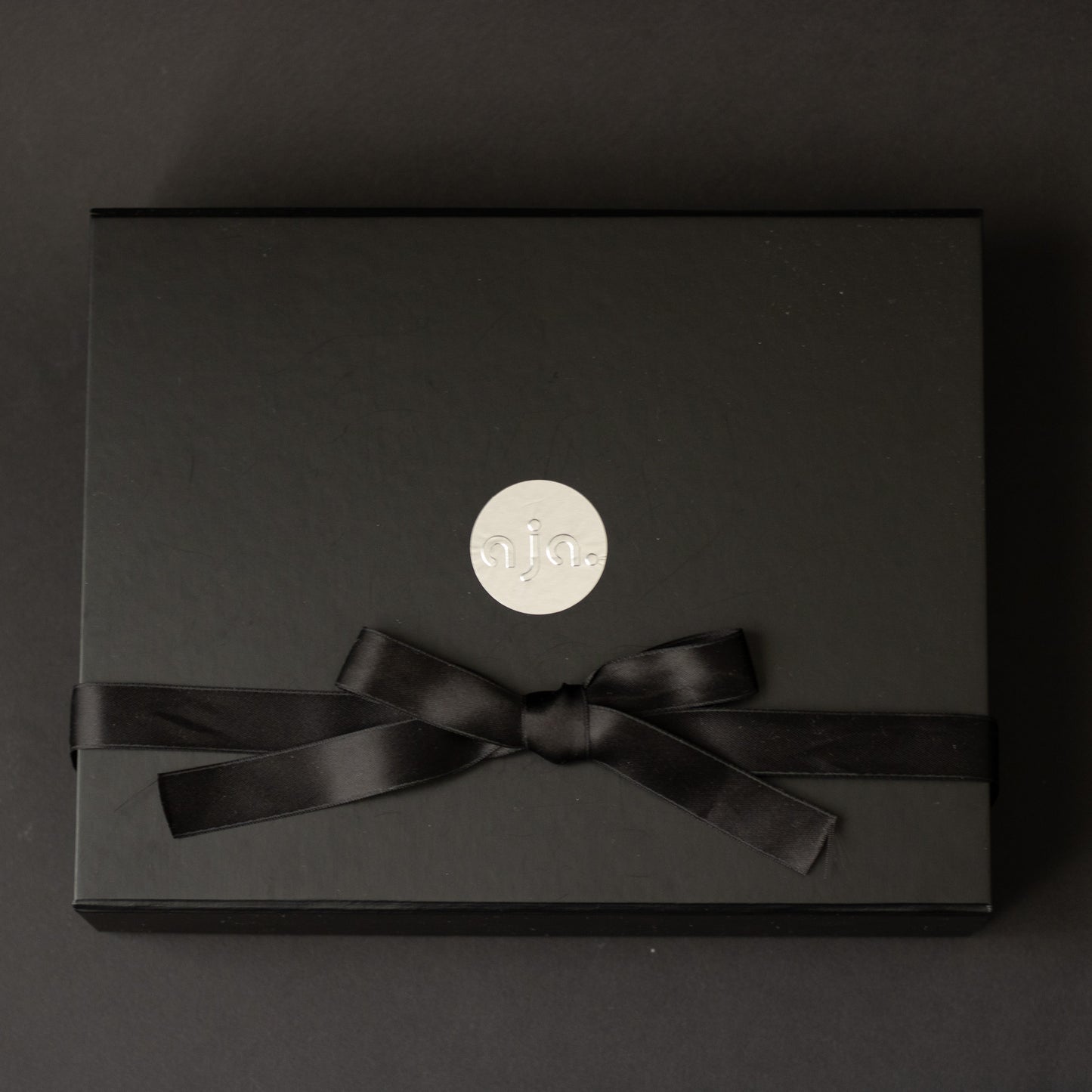 Black gift box with a black ribbon and 'aja' logo on a dark background

