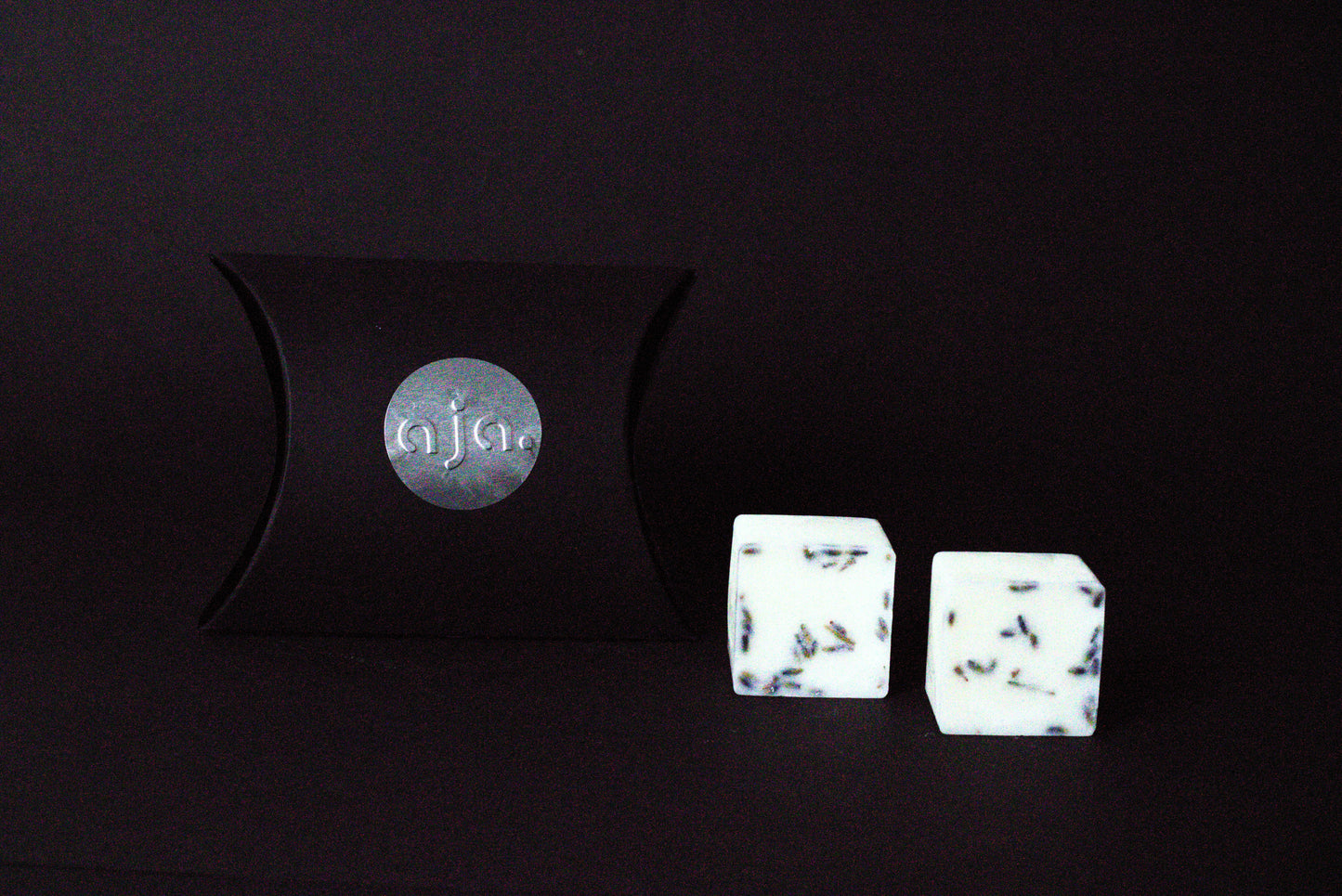 A black pillow box with a circular logo beside two  lavender botanical wax melt cubes on a dark background, conveying a minimalist and elegant vibe.