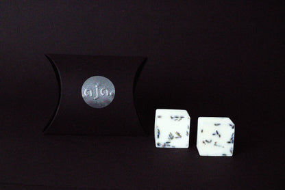 A black pillow box with a circular logo beside two  lavender botanical wax melt cubes on a dark background, conveying a minimalist and elegant vibe.