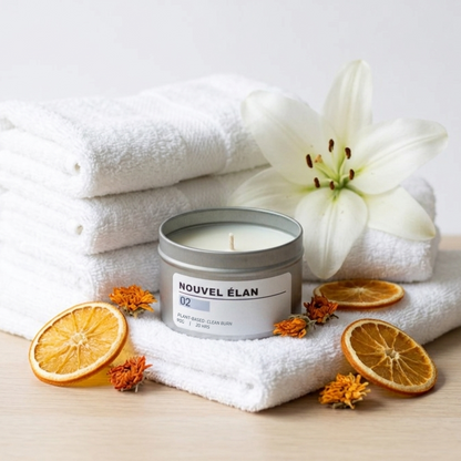 Candle labeled 'Nouvel Elan' on a stack of white towels with orange slices and a white flower.